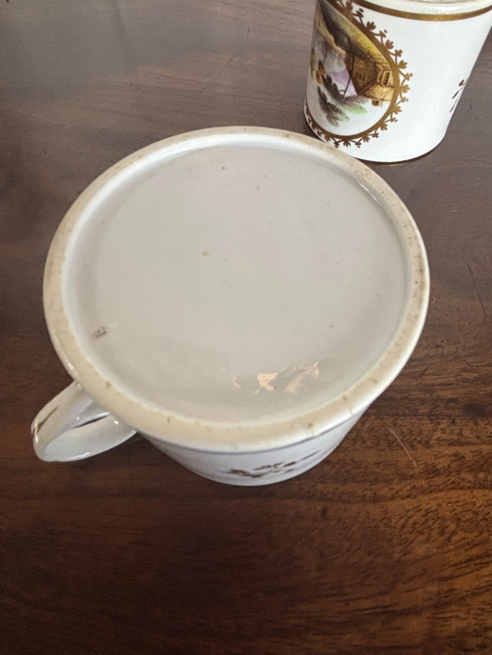 A pair of cylindrical white porcelain coffee cans decorated with hand-painted landscape vignettes enclosed in ornate gilded oval frames. View X