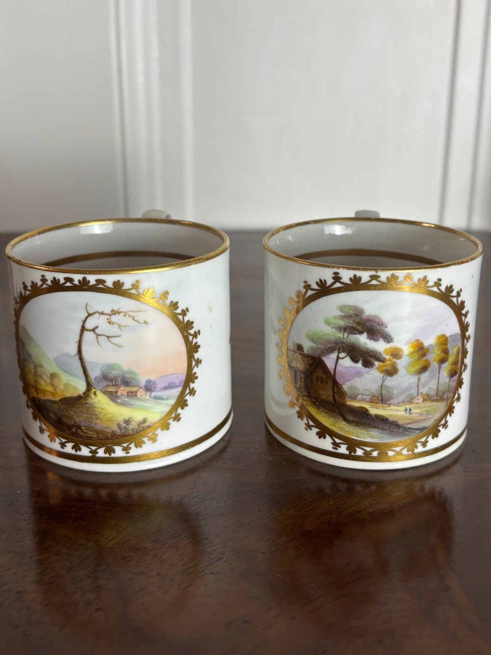 A pair of cylindrical white porcelain coffee cans decorated with hand-painted landscape vignettes enclosed in ornate gilded oval frames.