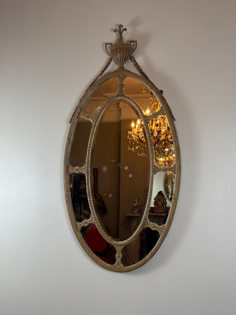 An antique oval wall mirror featuring a segmented border glass frame adorned with a classical top urn finial and carved swags. View III