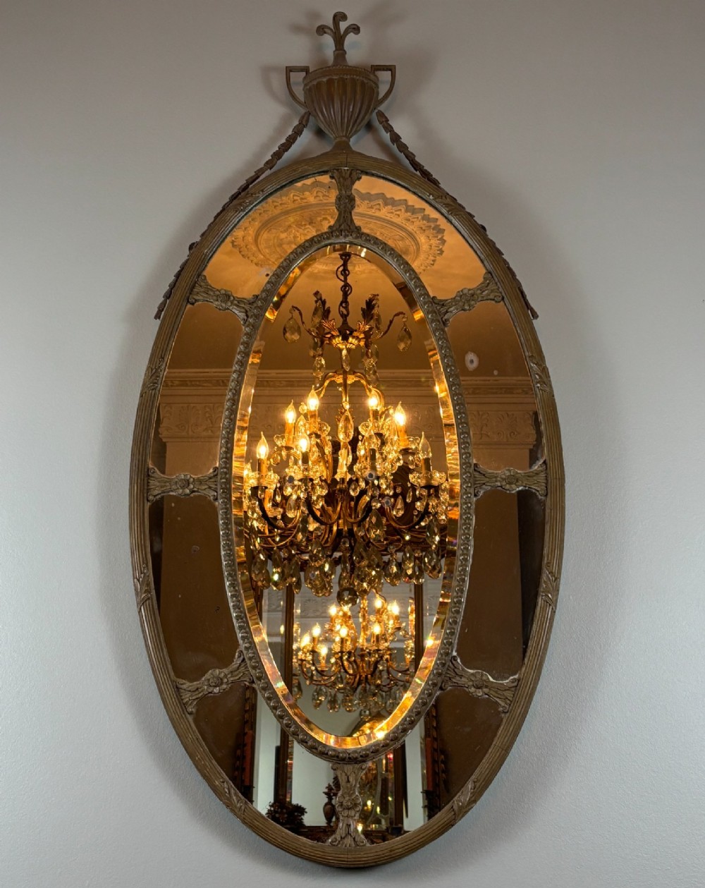 An antique oval wall mirror featuring a segmented border glass frame adorned with a classical top urn finial and carved swags.