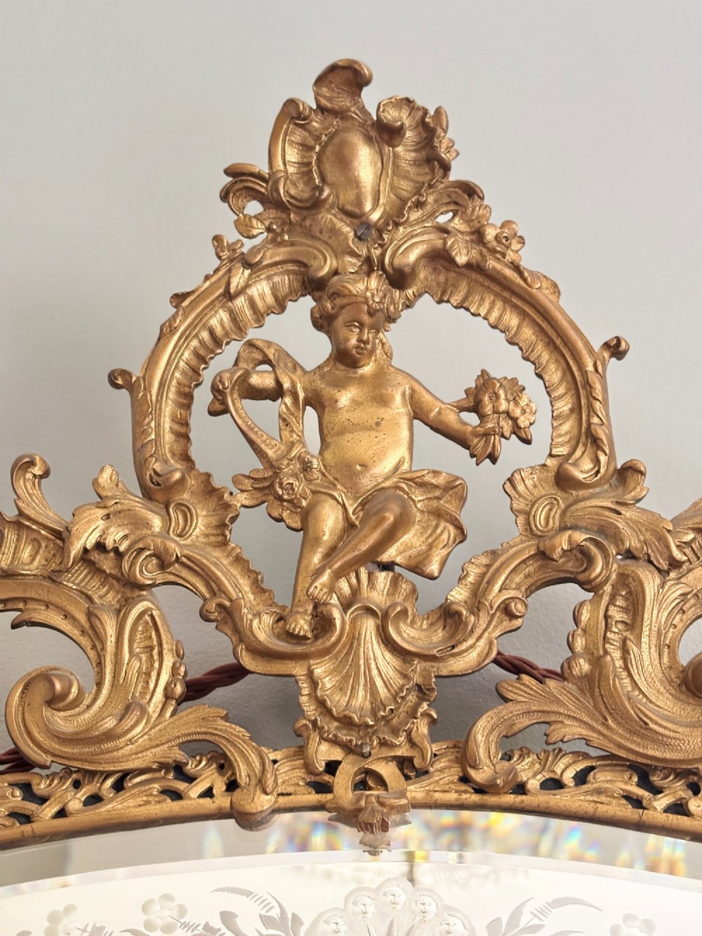 An ornate French Louis XV style gilt bronze wall mirror featuring etched glass borders, C-scroll framework, and a cherub cresting. View V