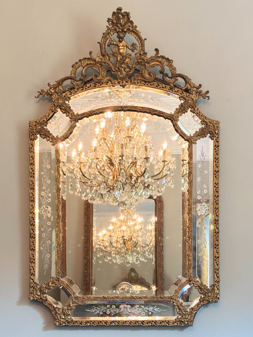 An ornate French Louis XV style gilt bronze wall mirror featuring etched glass borders, C-scroll framework, and a cherub cresting.