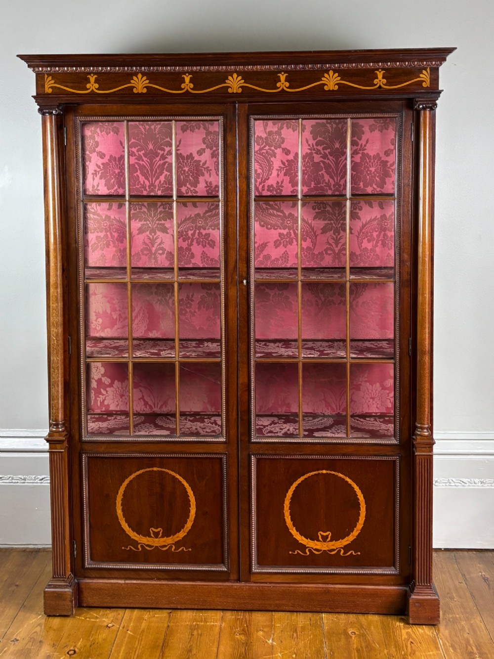A tall Georgian rosewood display cabinet featuring two glazed doors lined with red damask fabric, flanked by columns and adorned with marquetry inlays.