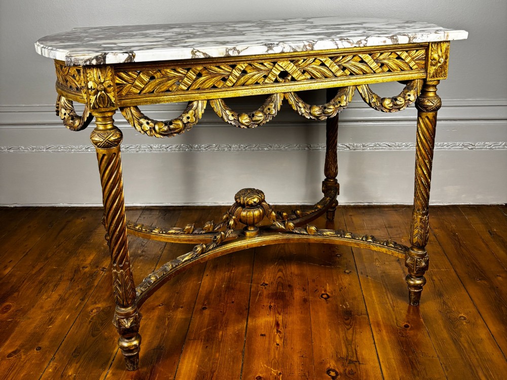 An antique French revival side table featuring a white carved marble top, a carved gold gilt frame with drapery swags, spiral-fluted legs, and a central cross stretcher.