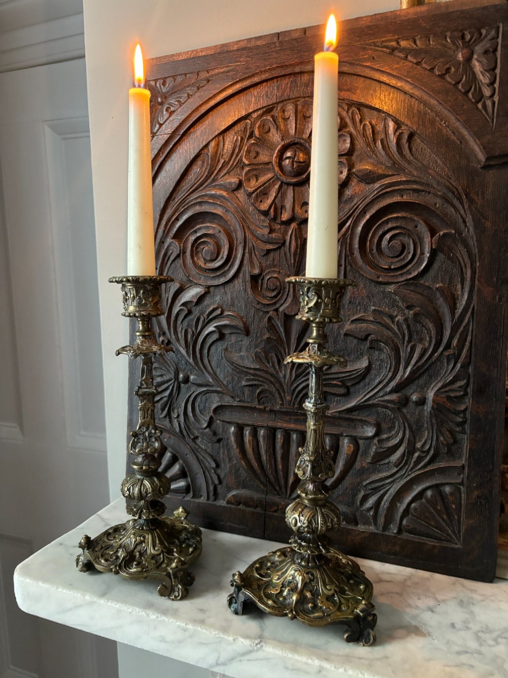 A pair of ornate 19th-century Regency brass candlesticks featuring detailed relief moulding and scrollwork bases holding lit candles. View V