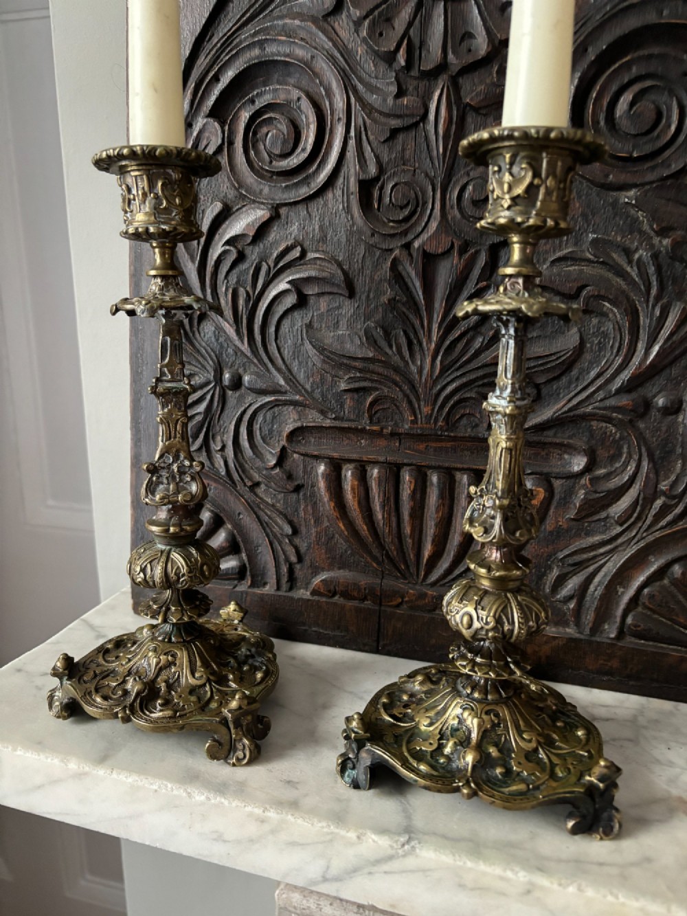 A pair of ornate 19th-century Regency brass candlesticks featuring detailed relief moulding and scrollwork bases holding lit candles. View IV