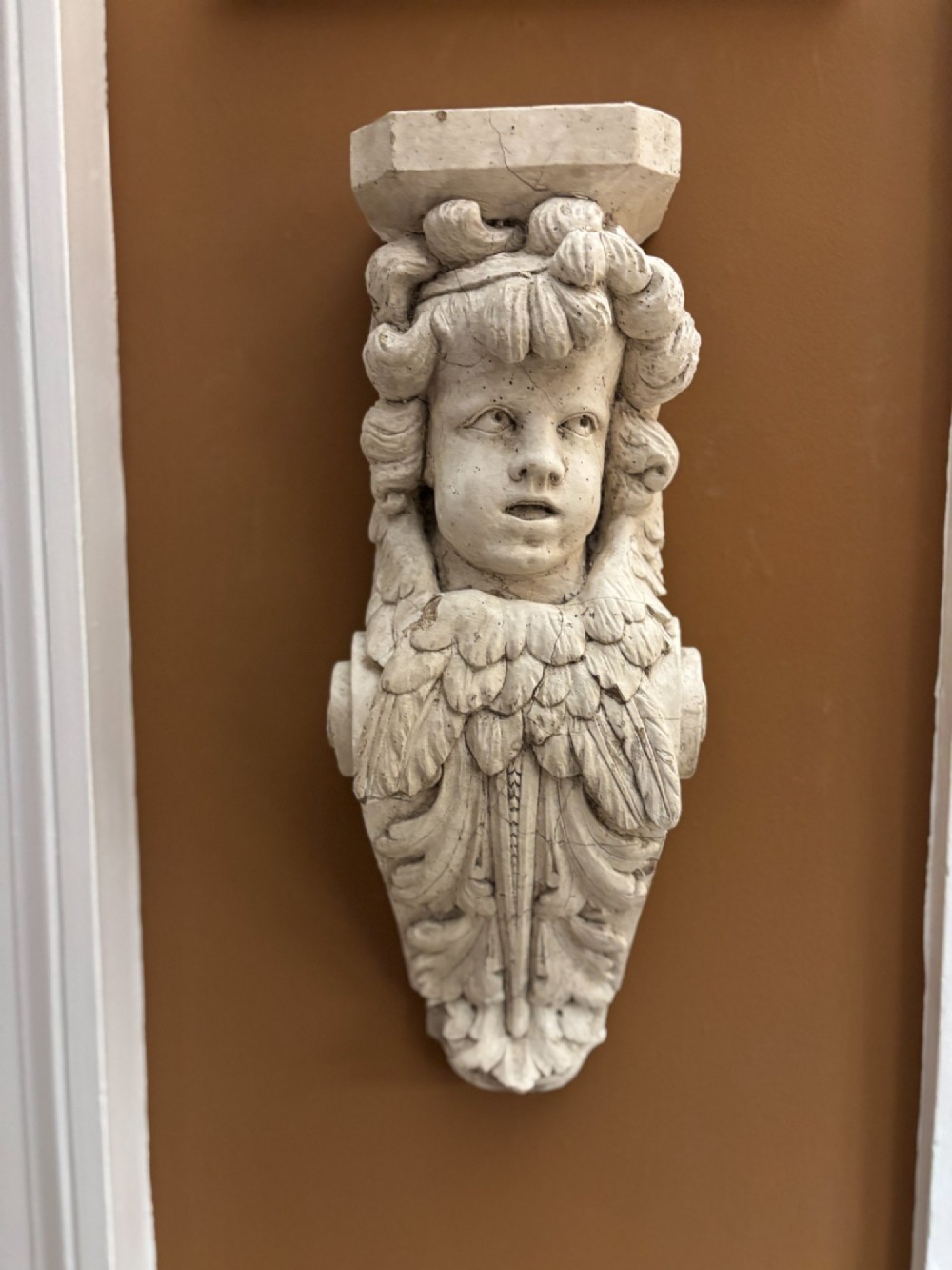 A 19th-century carved off-white architectural wall corbel featuring a winged putto head above acanthus leaf details. View VII