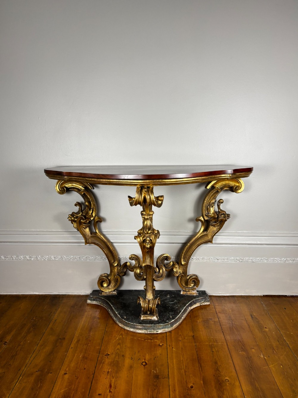 An 18th-century Venetian console table featuring intricate carved giltwood legs with scroll motifs resting on a shaped dark base beneath a polished dark timber top. View IX