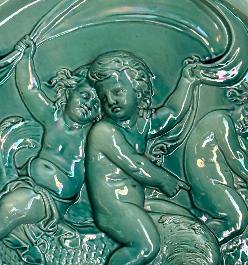 A circular teal ceramic wall plaque featuring a relief moulding of three cherubs riding a sea creature. View V