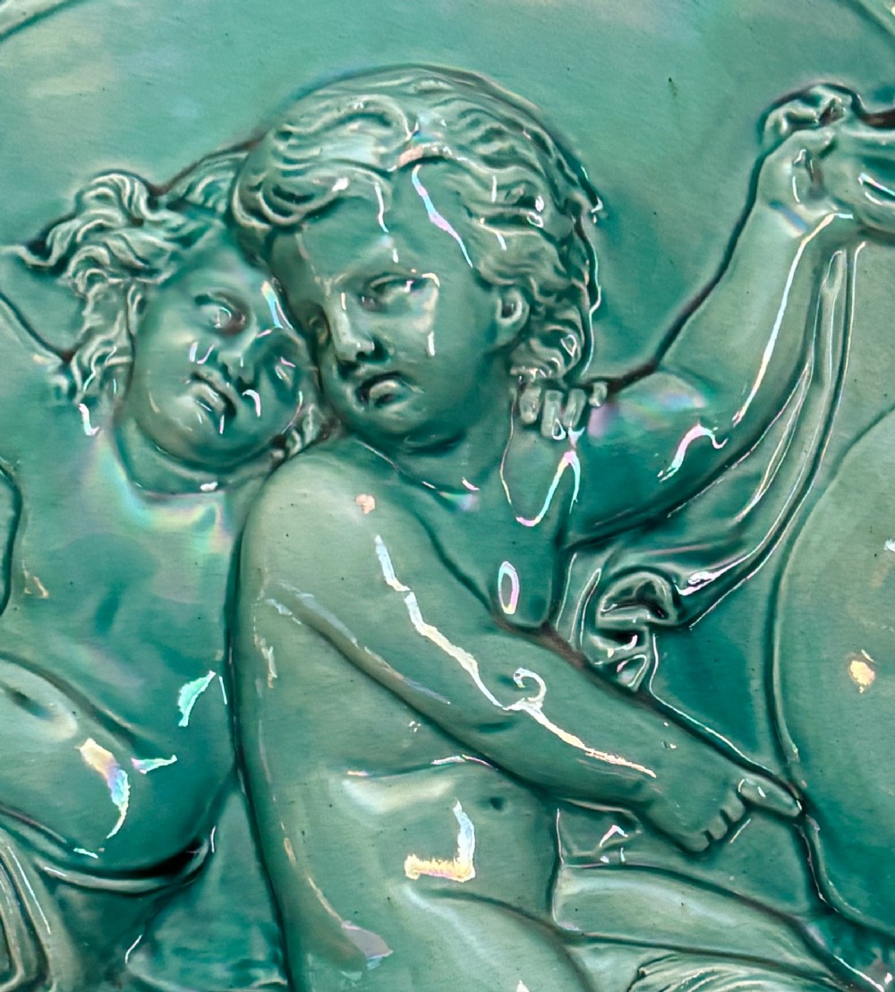 A circular teal ceramic wall plaque featuring a relief moulding of three cherubs riding a sea creature. View VII