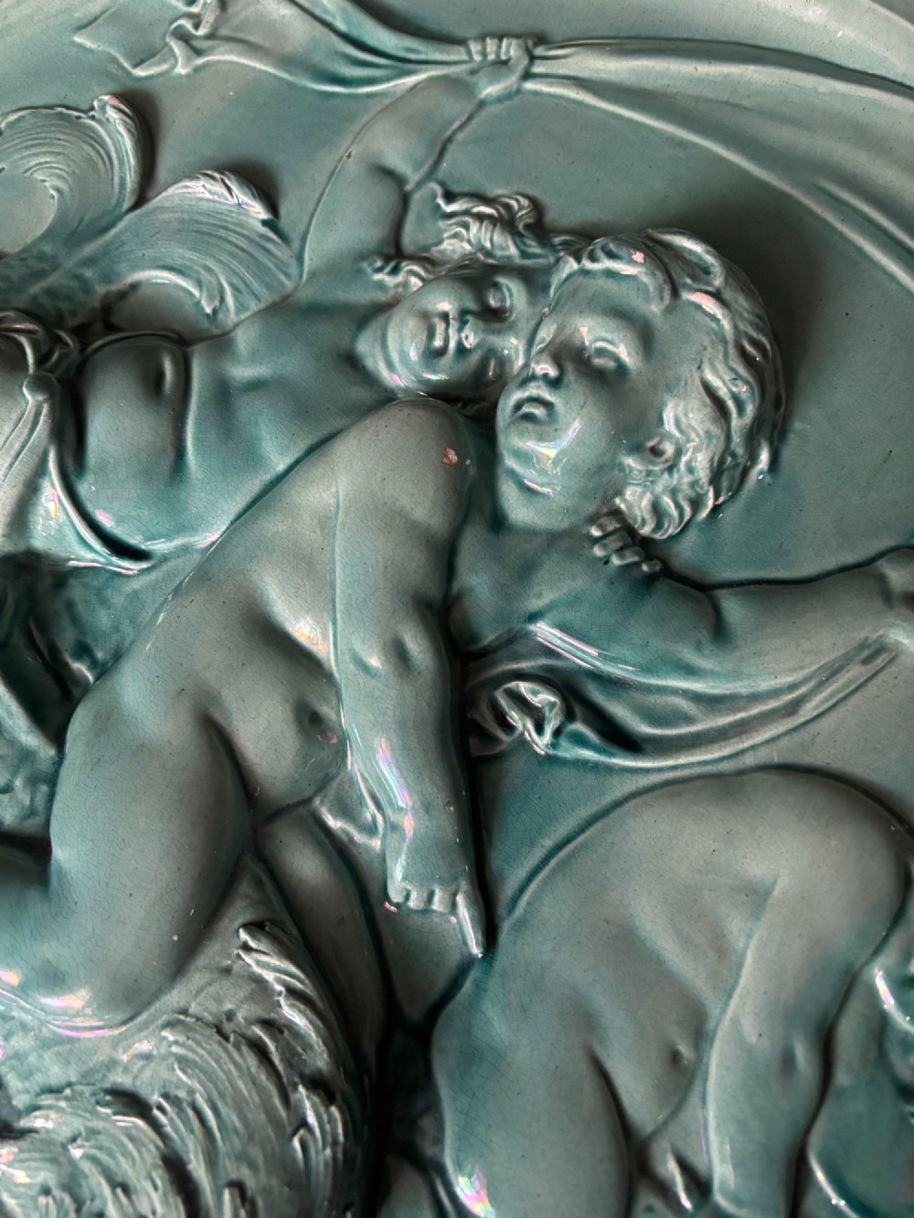 A circular teal ceramic wall plaque featuring a relief moulding of three cherubs riding a sea creature. View X