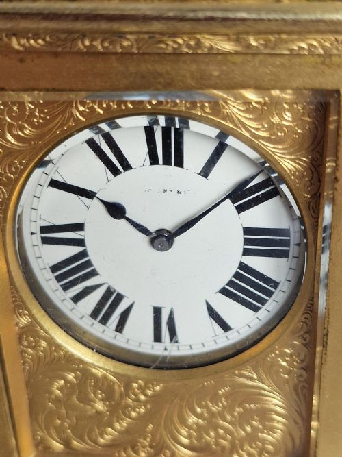 fig 1. A rectangular gilt brass carriage clock featuring elaborate foliate scroll engraving, a top carrying handle, glass panels, and a white enamel dial with Roman numerals.