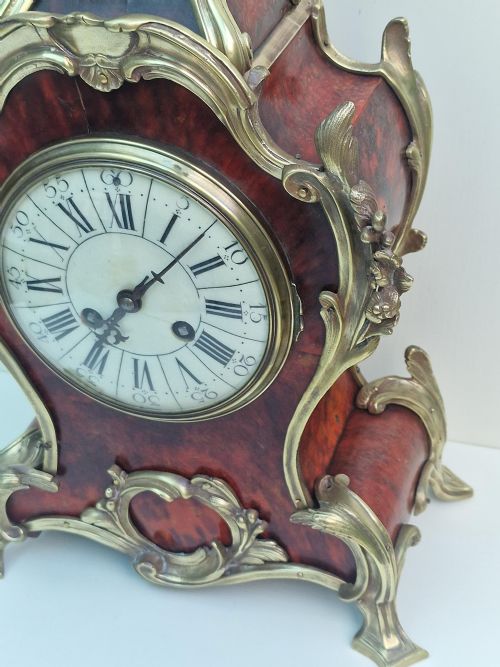 fig 1. A French Louis XV style tortoiseshell and ormolu mantel clock featuring a curved frame, foliage mounts, and a white enamel dial with Roman numerals.