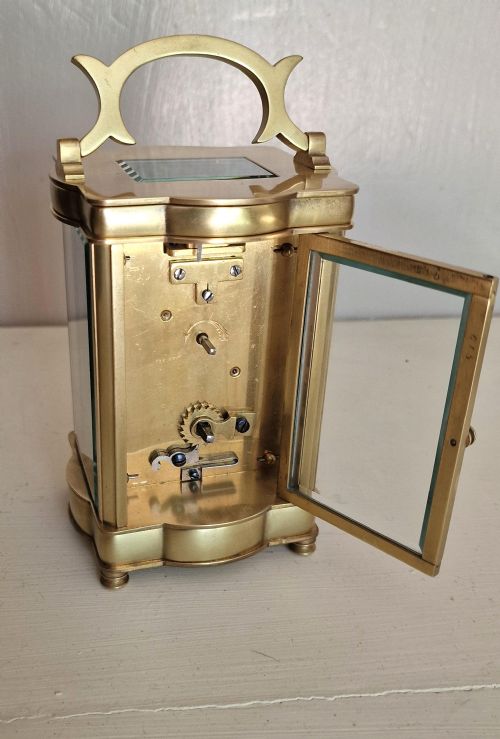 fig 1. A brass serpentine carriage clock featuring a white enamel dial with Roman numerals, bevelled glass panels, and an arched carrying handle.
