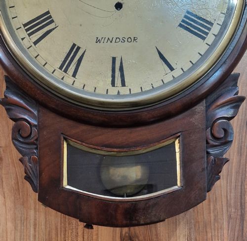 fig 1. An antique mahogany drop-dial wall clock featuring a painted dial with Roman numerals signed H. Lamb Windsor, a circular brass bezel, carved scroll ears, and a lower pendulum aperture.