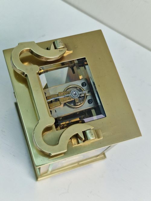 fig 1. A rectangular French brass carriage clock with bevelled glass panels, a white enamel dial with black Roman numerals, and a shaped carrying handle.