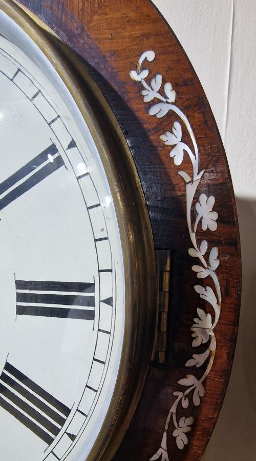fig 2. A Victorian drop-dial fusee wall clock featuring a white dial with Roman numerals, a dark wood case adorned with mother-of-pearl floral inlay, and a brass pendulum visible through a glass window.