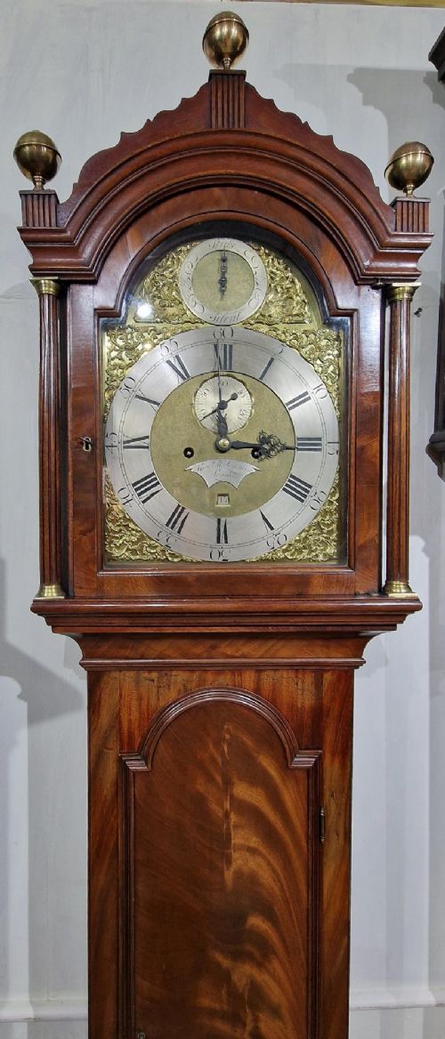 A tall Georgian flame mahogany longcase clock featuring an arched hood with brass finials, reeded columns, and a brass and silvered dial. View V