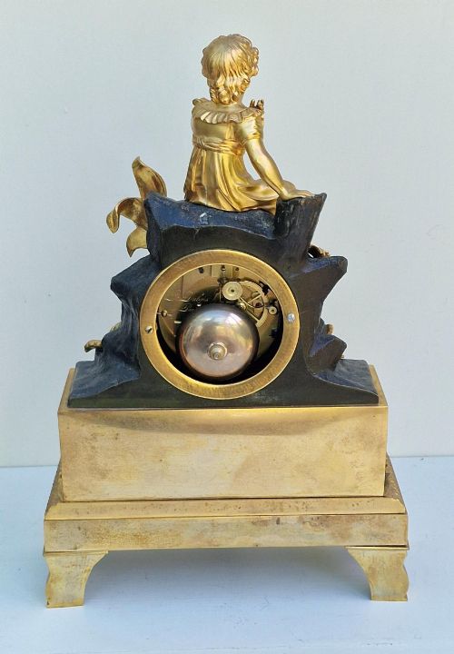 fig 1. A 19th-century French gilt bronze and patinated bronze mantel clock featuring a seated gilded child figure atop a dark rock-form casing with Roman numerals.