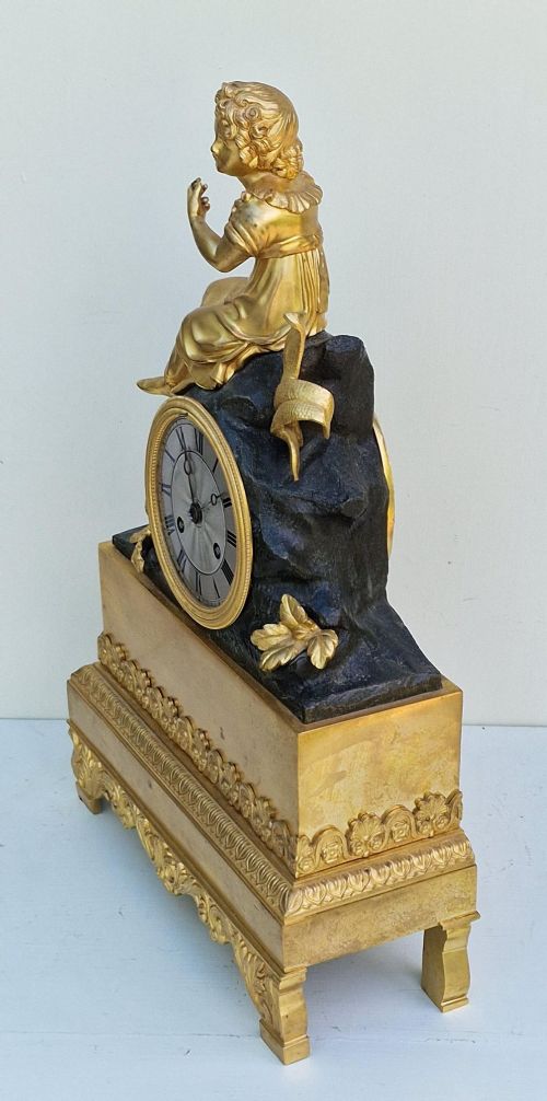 A 19th-century French gilt bronze and patinated bronze mantel clock featuring a seated gilded child figure atop a dark rock-form casing with Roman numerals. View VI