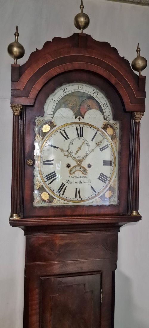 An antique Georgian flame mahogany longcase clock with three brass finials, an arched hood, fluted columns, and a painted dial displaying a moonphase mechanism. View VI