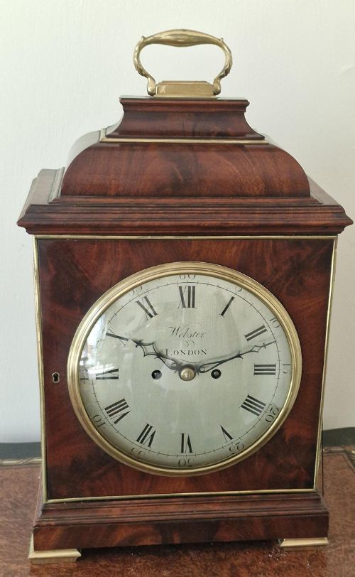 fig 1. A Georgian flame mahogany bracket clock featuring a brass carrying handle, circular white dial with Roman numerals, and brass-backed side frets.