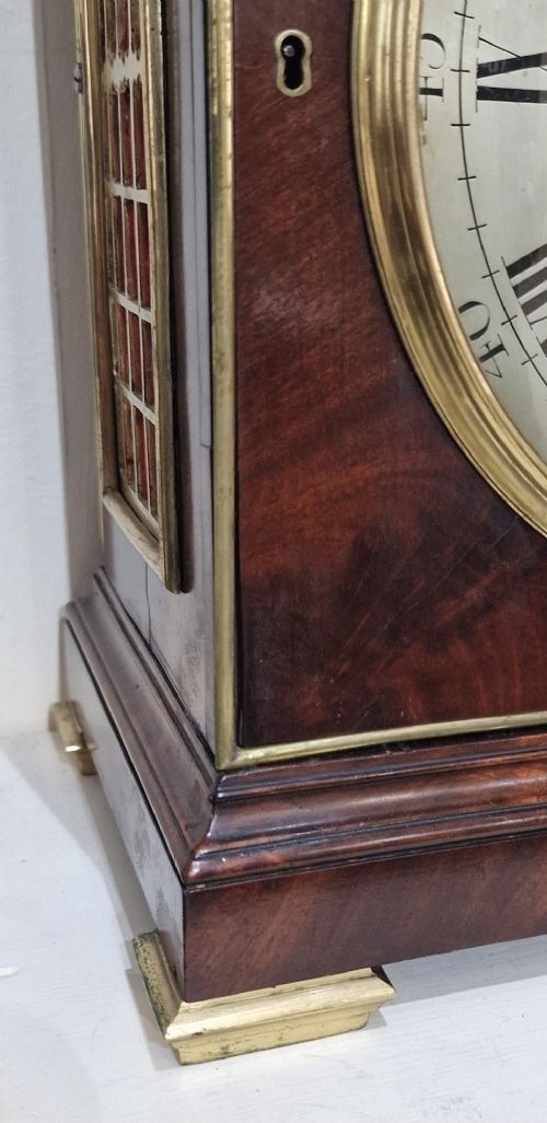 A Georgian flame mahogany bracket clock featuring a brass carrying handle, circular white dial with Roman numerals, and brass-backed side frets. View V