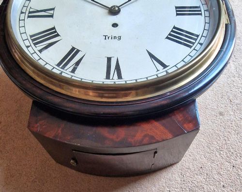 fig 1. A circular antique mahogany dial wall clock with a brass bezel, off-white face featuring Roman numerals, and black lettering reading Clarke Tring.