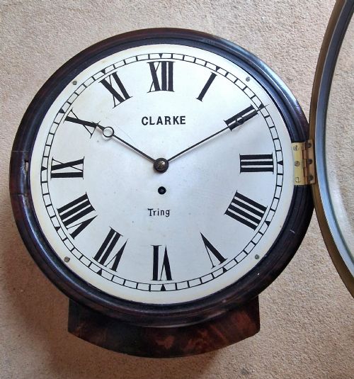 fig 2. A circular antique mahogany dial wall clock with a brass bezel, off-white face featuring Roman numerals, and black lettering reading Clarke Tring.