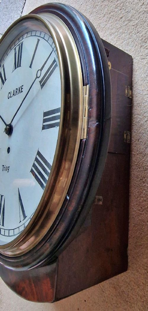 A circular antique mahogany dial wall clock with a brass bezel, off-white face featuring Roman numerals, and black lettering reading Clarke Tring. View V