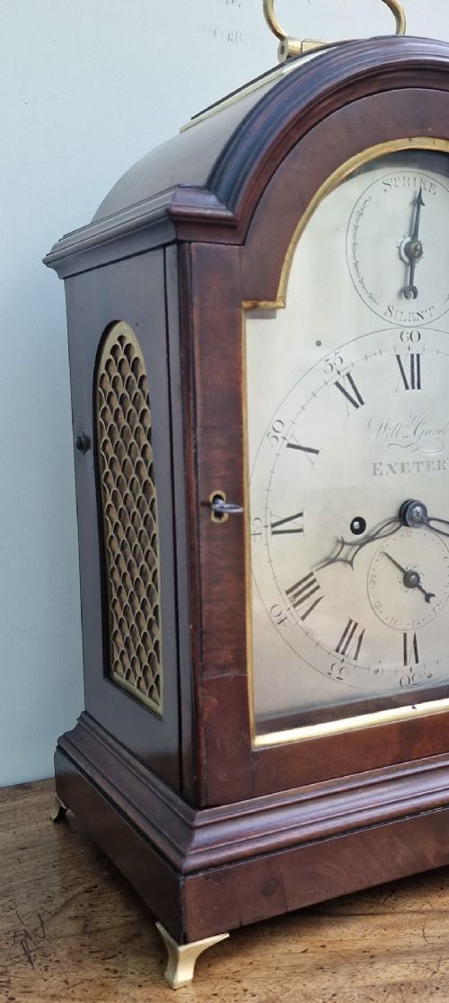 fig 2. A Georgian mahogany bracket clock featuring an arched top with a brass carrying handle, silvered engraved dial with Roman numerals, and brass bracket feet.