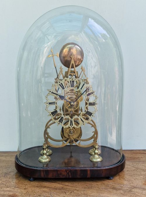 fig 1. A Victorian brass skeleton clock with an exposed mechanical movement and top strike bell, housed under a clear glass dome on an oval mahogany base.