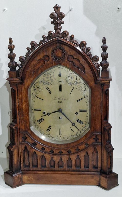 fig 1. A carved oak Gothic Revival bracket clock featuring pointed arch cresting, turned finials, blind tracery details, and an engraved brass dial with Roman numerals.