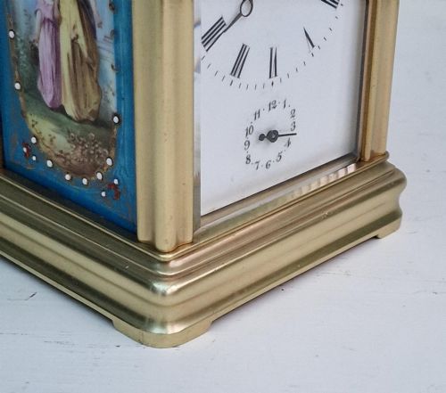 fig 1. A brass carriage clock featuring a hinged top handle, a glass viewing window above, a white enamel dial with Roman numerals, and side panels decorated with painted porcelain figurative scenes.
