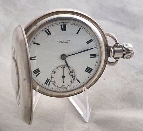 fig 1. A polished silver half hunter pocket watch displaying an engraved outer ring with black Roman numerals surrounding a central circular glass window.