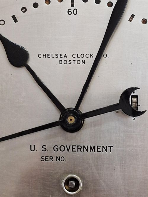 fig 1. A circular black maritime wall clock featuring a twenty-four-hour dial face marked U.S. Government and Chelsea Clock Co. Boston.