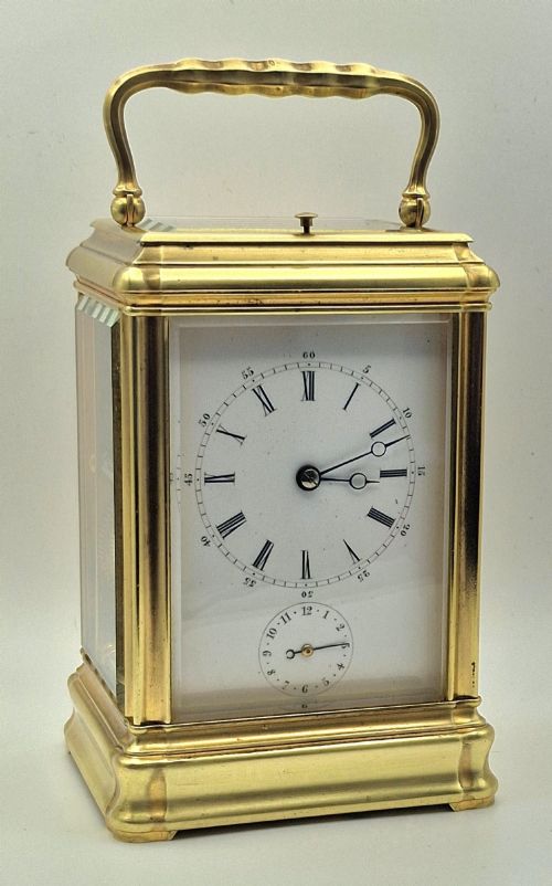 fig 1. A brass carriage clock featuring a gorge style case, rectangular glass panels, a top carrying handle, a white enamel dial with Roman numerals, an accompanying key, and a red fitted travel case.
