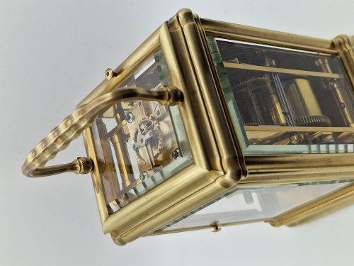 fig 2. A brass carriage clock featuring a gorge style case, rectangular glass panels, a top carrying handle, a white enamel dial with Roman numerals, an accompanying key, and a red fitted travel case.