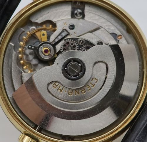 A 1957 Eterna-Matic 14K gold wristwatch with an off-white dial, date display window at three oclock, gold baton indices, and a black leather strap. View IX