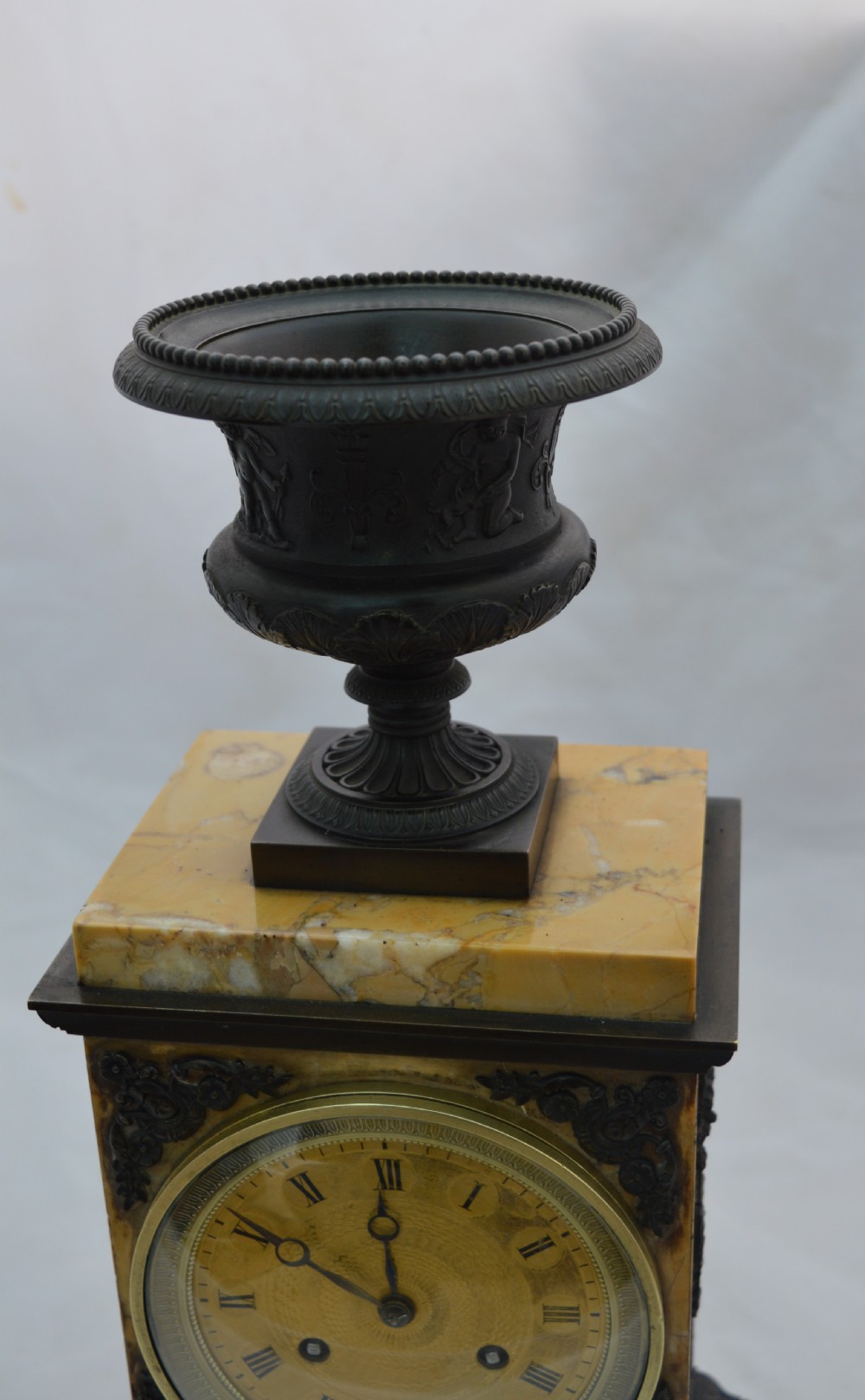 A French Empire mantel clock featuring a yellow marble rectangular body surmounted by a dark patinated bronze classical urn and housing a brass dial with Roman numerals. View III