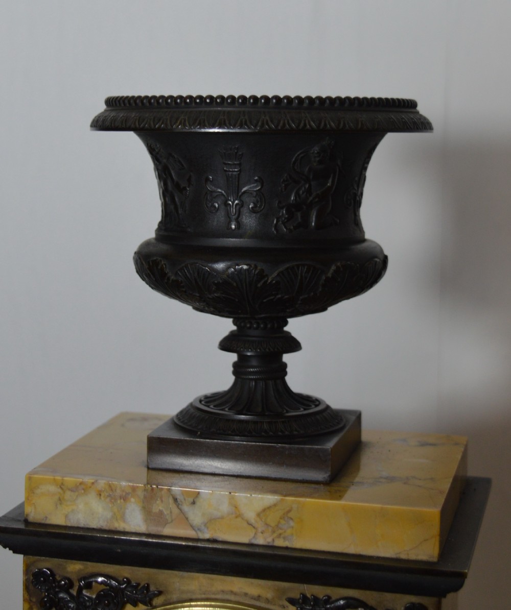 A French Empire mantel clock featuring a yellow marble rectangular body surmounted by a dark patinated bronze classical urn and housing a brass dial with Roman numerals. View IV