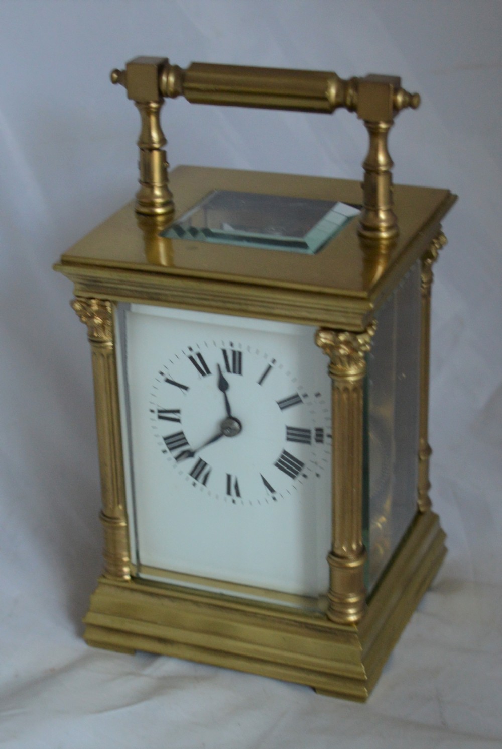 An antique brass carriage clock featuring Corinthian column corner supports, clear bevelled glass side panels, a top carrying handle, and a white enamel dial with black Roman numerals.