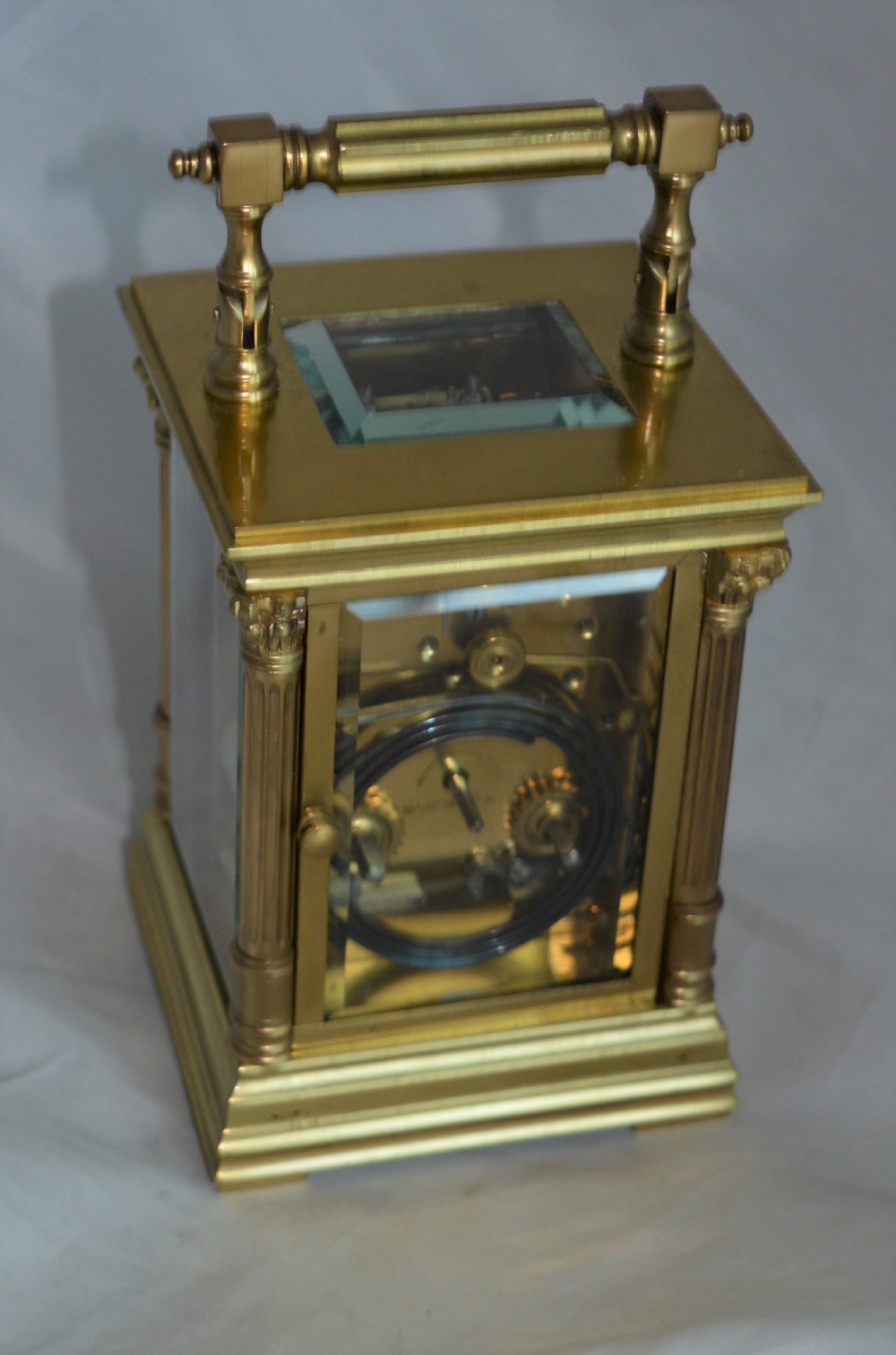 An antique brass carriage clock featuring Corinthian column corner supports, clear bevelled glass side panels, a top carrying handle, and a white enamel dial with black Roman numerals. View II