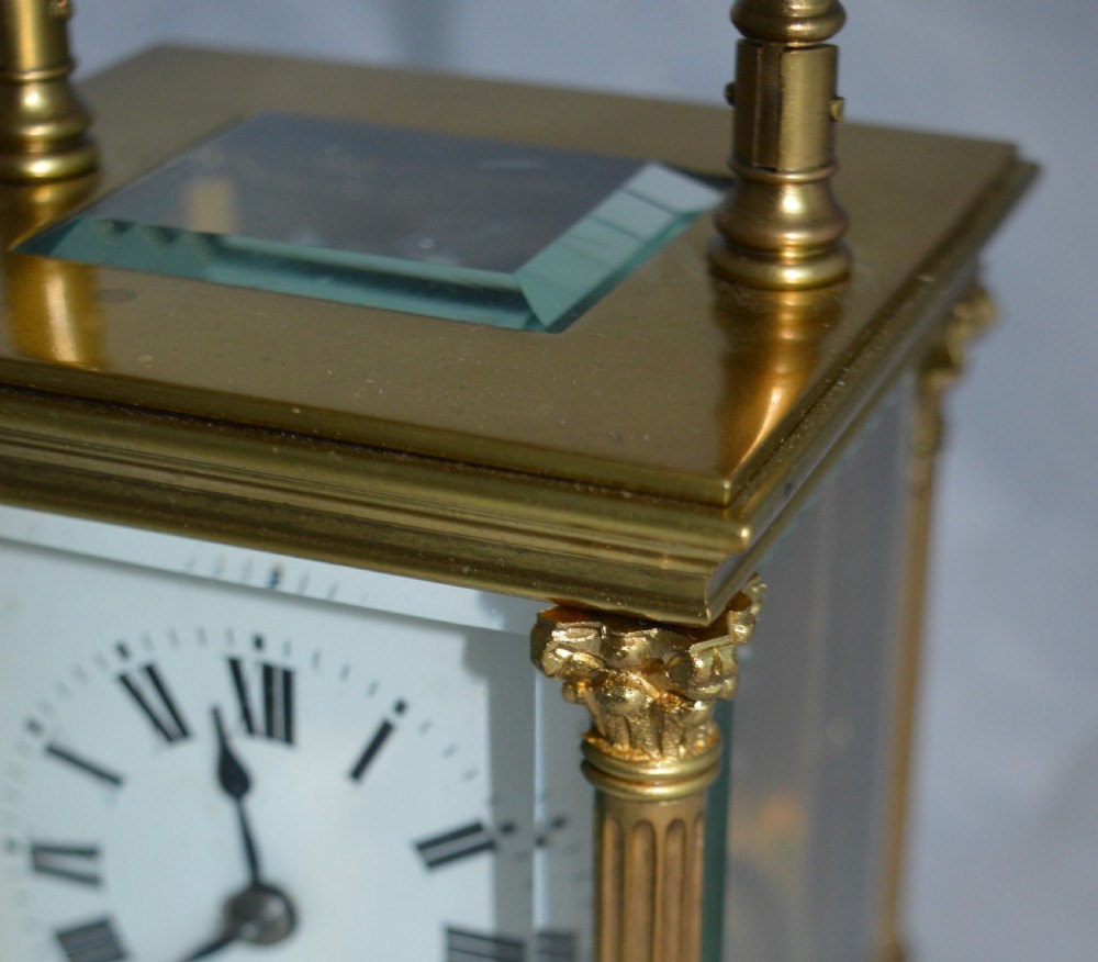 An antique brass carriage clock featuring Corinthian column corner supports, clear bevelled glass side panels, a top carrying handle, and a white enamel dial with black Roman numerals. View III