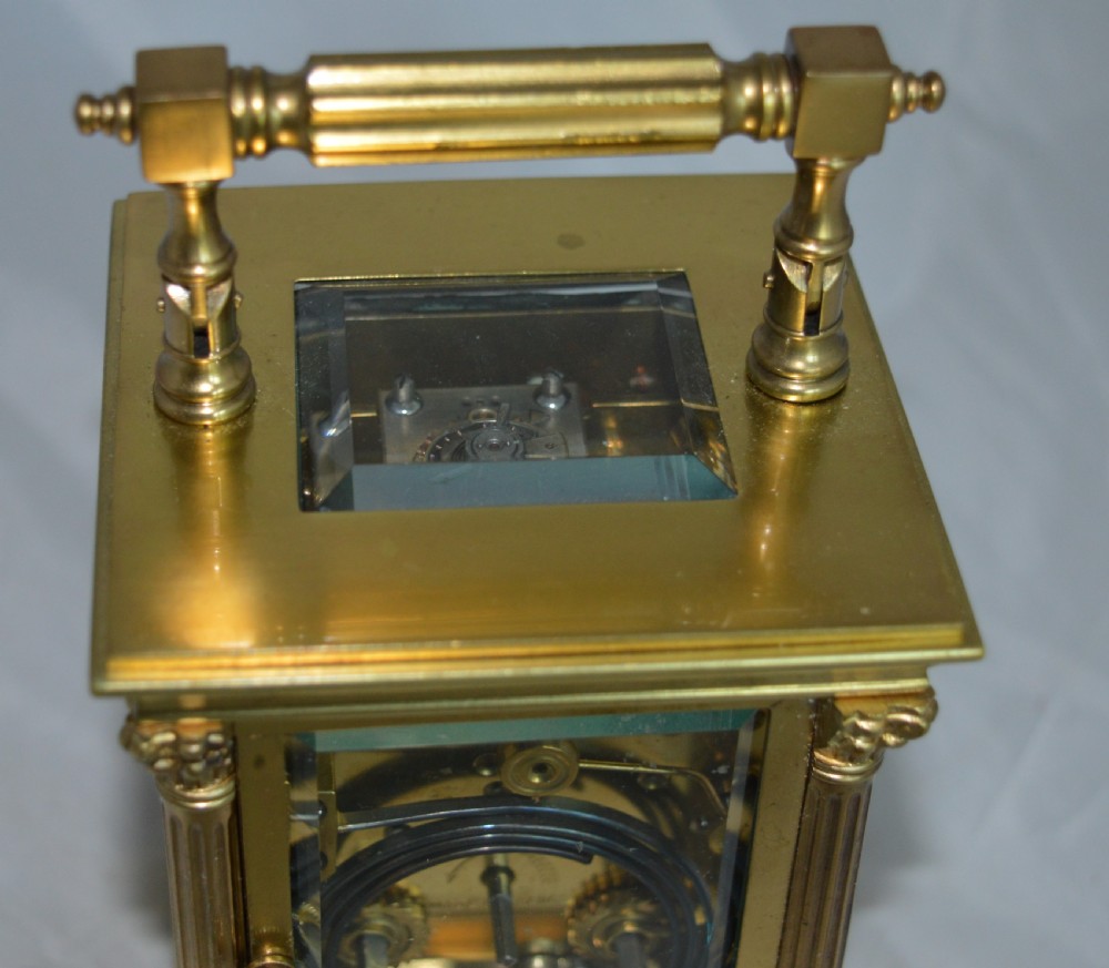 An antique brass carriage clock featuring Corinthian column corner supports, clear bevelled glass side panels, a top carrying handle, and a white enamel dial with black Roman numerals. View IV