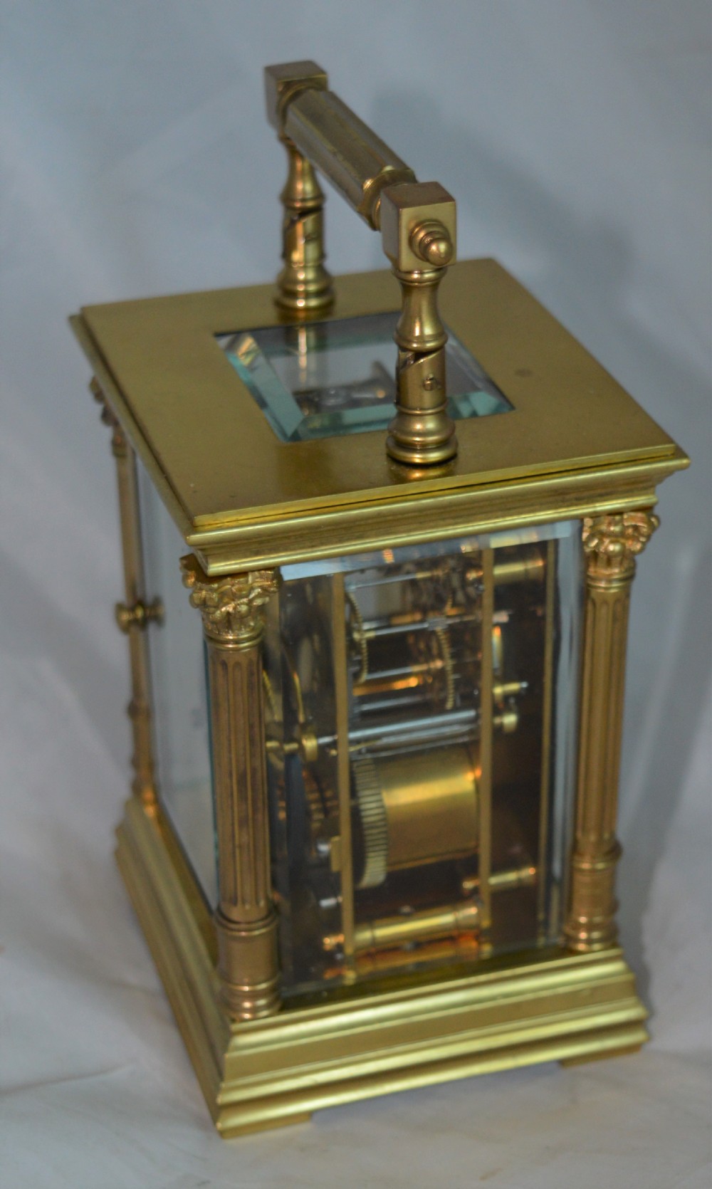 An antique brass carriage clock featuring Corinthian column corner supports, clear bevelled glass side panels, a top carrying handle, and a white enamel dial with black Roman numerals. View V