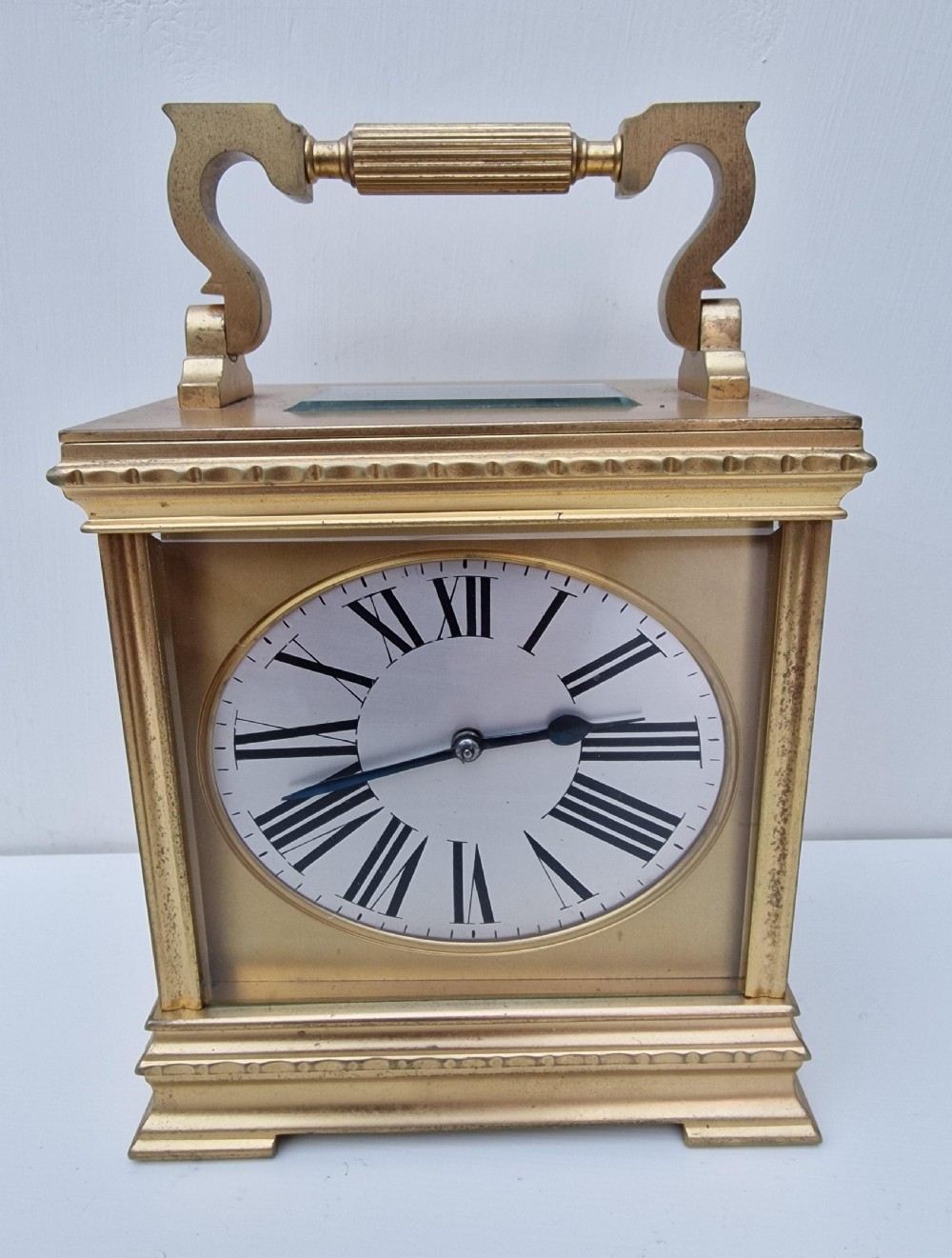 A brass carriage clock featuring an oval dial face with black Roman numerals and a decorative scrolled handle.