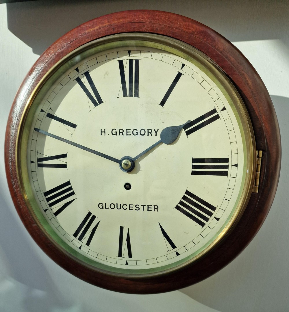 A circular antique mahogany wall dial clock featuring a brass bezel, off-white face with Roman numerals, black hands, and the makers mark H. Gregory Gloucester.