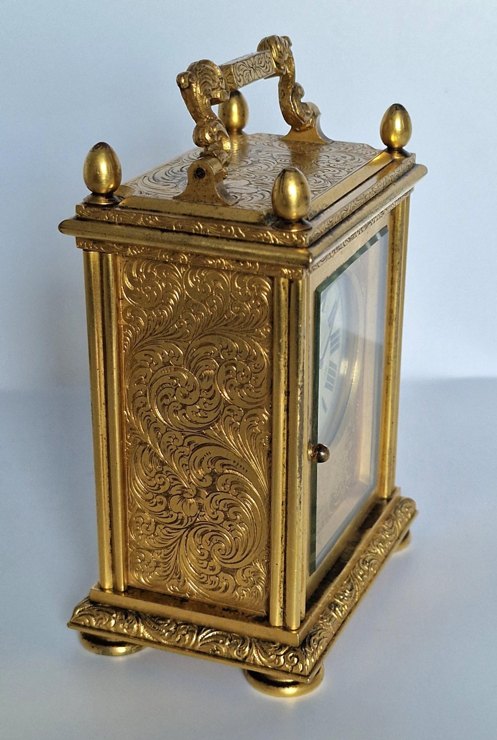 A rectangular gilt brass carriage clock featuring elaborate foliate scroll engraving, a top carrying handle, glass panels, and a white enamel dial with Roman numerals. View III