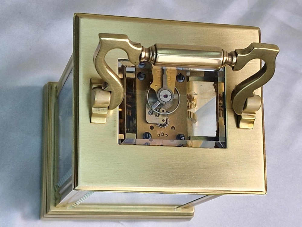 A rectangular brass carriage clock featuring a hinged top carrying handle, clear glass panels, and a white enamel dial with black Roman numerals. View II